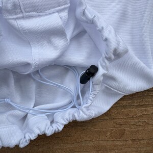 May include: A white mesh garment featuring a gathered hem with a white drawstring and a black plastic cord lock. The fabric has a textured, grid-like pattern, and a portion of a blue horizontal stripe is visible at the top edge of the item.