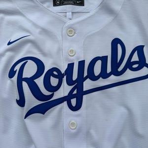 May include: A white button-down baseball jersey featuring the word Royals in blue script across the chest. The garment includes a small blue Nike swoosh logo on the upper right chest and a black Nike label at the inner neckline with size M.