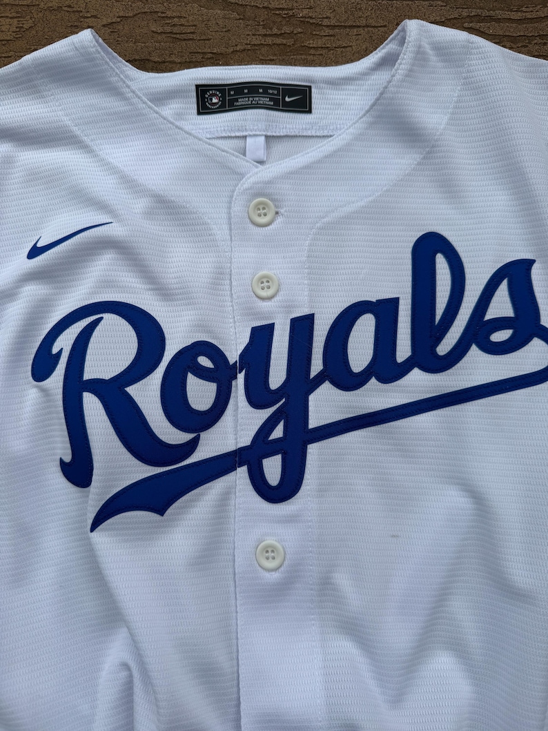 May include: A white button-down baseball jersey featuring the word Royals in blue script across the chest. The garment includes a small blue Nike swoosh logo on the upper right chest and a black Nike label at the inner neckline with size M.