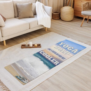 May include: A yoga mat with a blue, white, and orange sunset background. The text "Yoga Dad" is in blue and white, and the text "Like a regular dad but cooler" is in white. The mat has a beach scene with waves and a pair of flip-flops.
