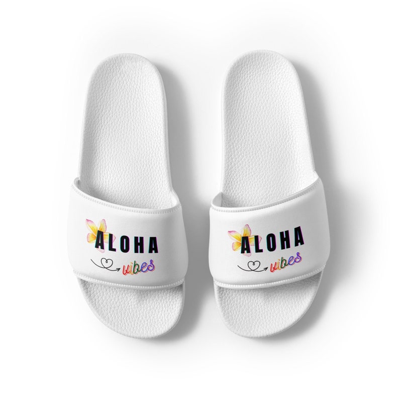 K&ouml;nnte beinhalten: Wei&szlig;e Rutschsandalen mit einem schwarzen "ALOHA vibes"-Aufdruck und einem gelben Blumendesign.