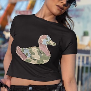 May include: Black crop top featuring a duck applique. The duck design is made of fabric with a camouflage pattern in shades of green, pink, and blue. The shirt has short sleeves and a round neckline.