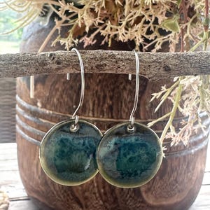 May include: A pair of round, ceramic drop earrings featuring a glossy, mottled glaze in shades of deep teal, forest green, and olive. The circular pendants are suspended from simple, curved silver-toned metal ear wires for a clean, polished look.