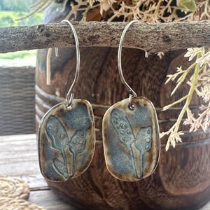 May include: A pair of drop earrings featuring rectangular ceramic charms with rounded edges. The charms have a glazed finish in shades of blue and brown, displaying a botanical print of leaves. The earrings hang from silver-toned metal hooks.
