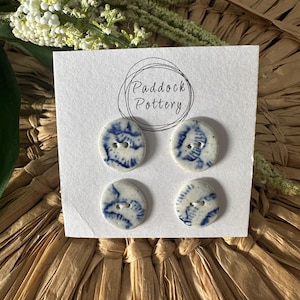 May include: Four ceramic buttons with a white base and blue floral designs are displayed on a white card. The card has the text "Paddock Pottery" in a circular design. The buttons are on a woven straw surface, with greenery in the background.