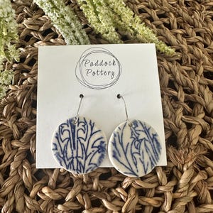 May include: Round, white ceramic earrings featuring a blue floral pattern. The earrings have silver-coloured hooks. The earrings are presented on a white card with the text "Paddock Pottery" in a circular design.