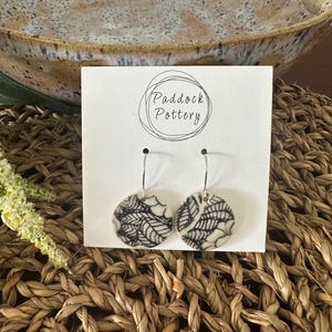 May include: Round ceramic earrings featuring a black and white spiderweb pattern. The earrings hang from silver-coloured hooks and are presented on a white card with the text "Paddock Pottery". A woven basket and ceramic bowl form the background.