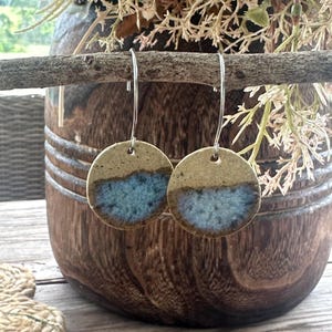 May include: A pair of circular ceramic earrings featuring a speckled tan base with a textured blue glaze across the bottom half. The earrings are suspended from silver-toned metal hooks, showcasing a handcrafted aesthetic with distinct colour variations.