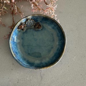 May include: A small, round, handmade ceramic dish with a glossy blue glaze. The rim has a golden-brown edge. Three raised floral designs are on the rim. The dish is on a grey surface, with pink floral branches in the background.