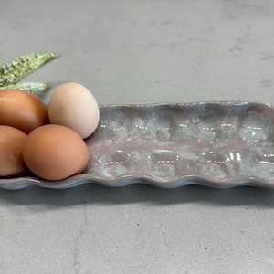 May include: A ceramic egg holder with a wavy edge, glazed in shades of blue and pink, holds four brown eggs and one white egg. The egg tray is designed to hold a dozen eggs. A decorative plant is in the background.