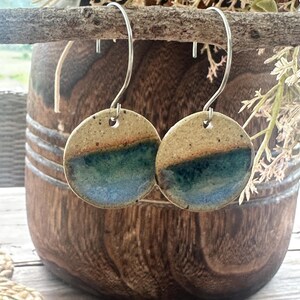 May include: A pair of round ceramic earrings featuring a speckled beige top half and a glossy blue-green glazed bottom half. The earrings hang from silver-toned metal hooks, showcasing a distinct colour transition across the circular clay surface.