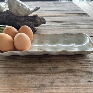May include: A ceramic egg tray with a wavy edge holds four brown eggs. The tray is a light beige colour with a slightly textured surface. The eggs are a warm brown colour. The tray is on a wooden surface.