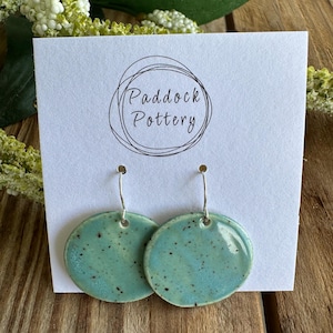 May include: Turquoise ceramic earrings with a speckled glaze. The round earrings hang from silver-coloured hooks. The earrings are displayed on a white card with the text "Paddock Pottery" in a circular design.