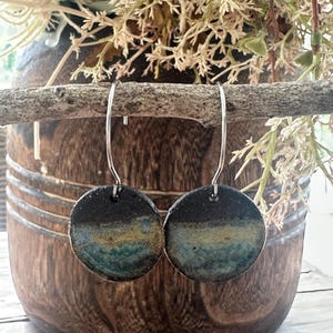 May include: A pair of circular earrings featuring a textured, multi-coloured glaze with gradients of black, deep blue, and earthy green. The round ceramic discs are suspended from thin, silver-toned metal hooks, creating a simple and balanced design.