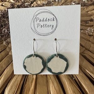May include: A pair of white ceramic earrings with a green glaze around the edges. The earrings are hanging from silver hooks. The earrings are on a white card with the text "Paddock Pottery" in black.