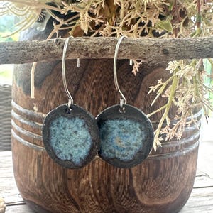 May include: A pair of circular earrings featuring a dark, textured outer rim and a speckled, light blue centre. The earrings are suspended from silver-toned metal hooks, showcasing a handcrafted appearance with a distinct contrast between the dark edges and the vibrant blue glaze finish.