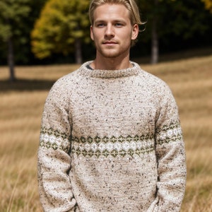 Op de afbeelding: Een beige trui met een groene en witte band met patroon rond de borst. De trui heeft een gespikkelde textuur.