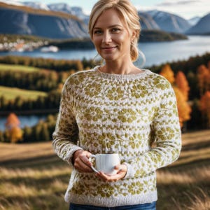 May include: A woman wearing a white and green patterned sweater with a white mug in her hands. She is standing in a field with a mountain range in the background.