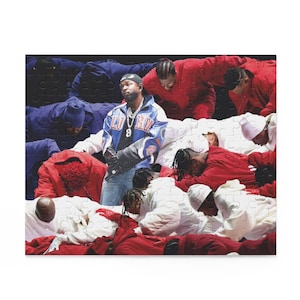 May include: A man wearing a blue and white jacket with a gold chain stands in front of a group of people wearing red and white clothing. The people are lying down in a circle around the man.