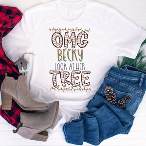 May include: White t-shirt featuring a Christmas-themed graphic that reads "OMG Becky Look at her Tree" with red and green striped letters and a string of colourful lights.