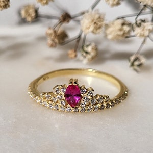 May include: A gold ring with a pink oval-shaped gemstone surrounded by smaller clear gemstones. The ring is designed in a crown shape.