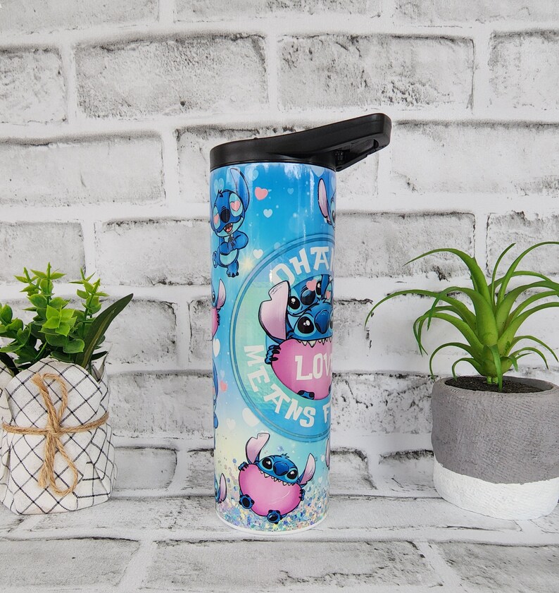 May include: A blue and pink stainless steel tumbler with a black lid. The tumbler features a repeating pattern of Stitch, a blue alien character from the Disney movie Lilo & Stitch, holding a pink heart. The text "Ohana Means Family" is printed on the tumbler.