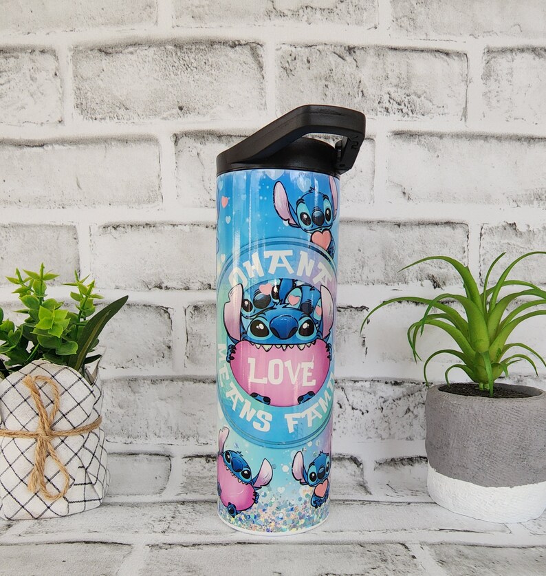 May include: A blue and pink stainless steel tumbler with a black lid and handle. The tumbler features a repeating pattern of Stitch, a blue alien character from the Disney movie Lilo & Stitch, holding a pink heart that says "Love" and a circle that says "Ohana Means Family".