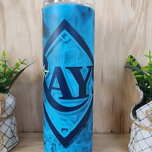 May include: A tall, insulated tumbler with a clear lid and straw. The tumbler features a vibrant blue design with the letters "AY" in a dark blue geometric shape. The background has a smoky, abstract pattern.