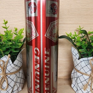K&ouml;nnte beinhalten: Ein Edelstahlbecher mit einem roten und silbernen Design. Der Becher zeigt ein sich wiederholendes Muster eines Footballhelms und das Wort "GENUINE".