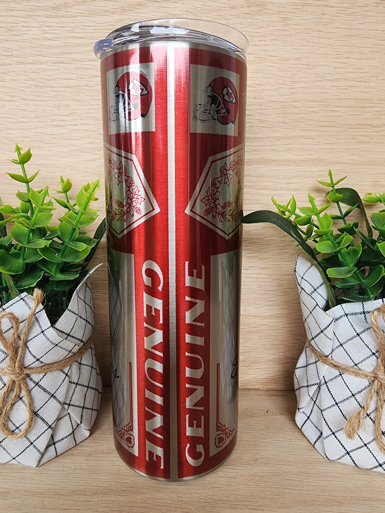 K&ouml;nnte beinhalten: Ein Edelstahlbecher mit einem roten und silbernen Design. Der Becher zeigt ein sich wiederholendes Muster eines Footballhelms und das Wort "GENUINE".
