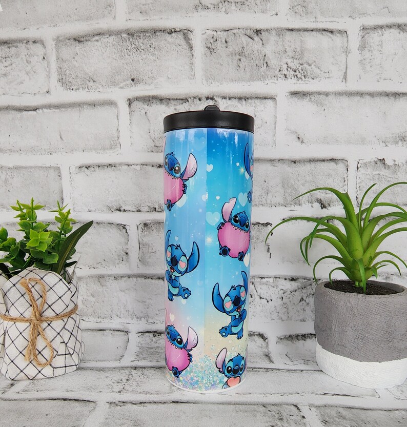 May include: A blue and pink stainless steel tumbler with a lid. The tumbler features a repeating pattern of a blue cartoon character with pink hearts.