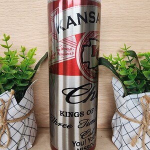 K&ouml;nnte beinhalten: Ein Edelstahlbecher mit einem roten und silbernen Design. Der Becher hat ein Etikett im Vintage-Stil mit dem Text "KANSAS" und "THE WORLD'S RENOWNED". Der Boden des Bechers tr&auml;gt den Text "KINGS OF", "Three Time", "Est", "YOU! YOU'RE", und "MISSOURI".