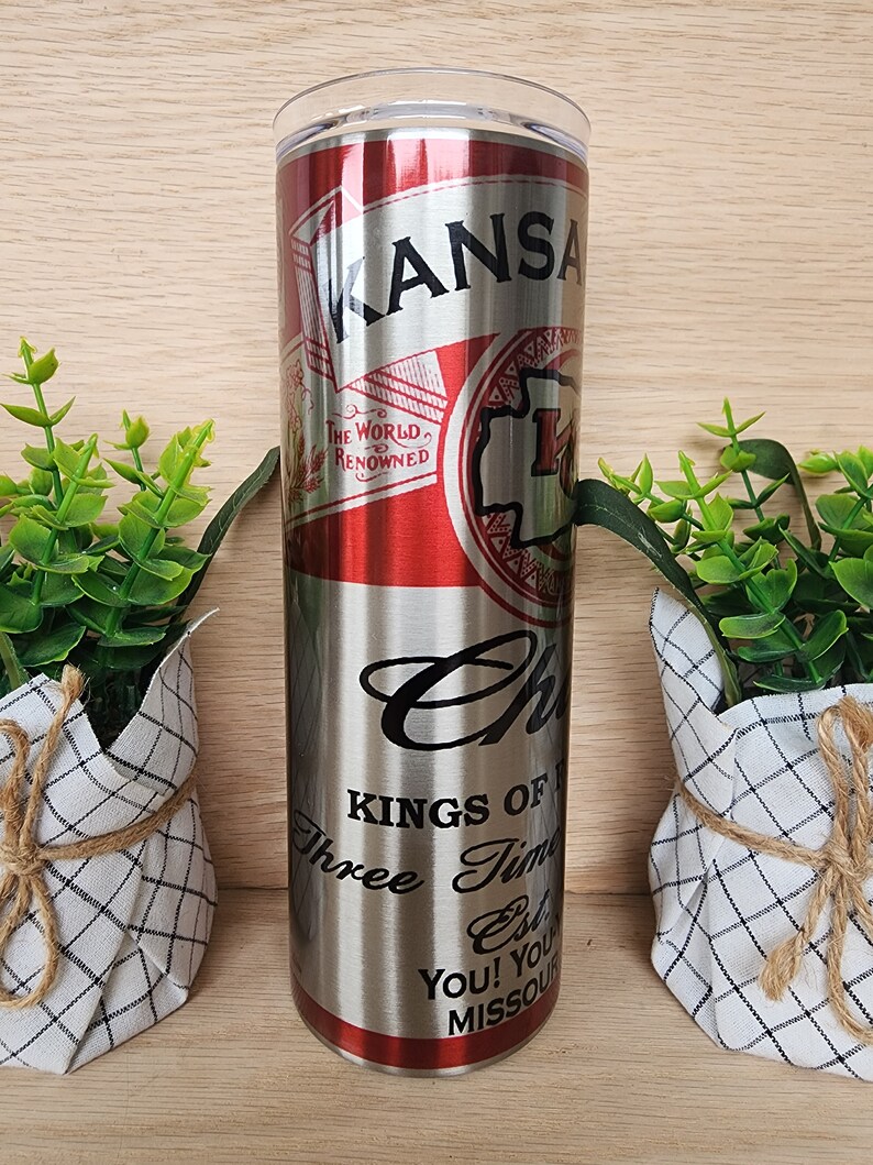 K&ouml;nnte beinhalten: Ein Edelstahlbecher mit einem roten und silbernen Design. Der Becher hat ein Etikett im Vintage-Stil mit dem Text "KANSAS" und "THE WORLD'S RENOWNED". Der Boden des Bechers tr&auml;gt den Text "KINGS OF", "Three Time", "Est", "YOU! YOU'RE", und "MISSOURI".