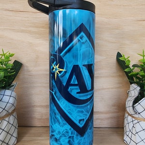 Puede incluir: Un vaso de acero inoxidable azul y negro con un logotipo de los Tampa Bay Rays y un diseño de llamas. El vaso tiene una tapa negra con una asa de transporte.