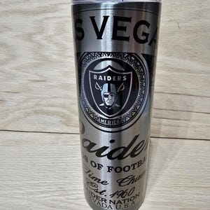Pode incluir: Um copo de aço inoxidável prateado com um logotipo preto dos Raiders e texto que diz "Las Vegas Raiders, Time of Football, Est. 1960, Raider Nation, Nevada U.S.A."