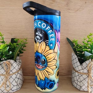 Puede incluir: Un vaso de acero inoxidable azul y amarillo con un dise&ntilde;o de personaje de dibujos animados. El personaje es Stitch de Lilo & Stitch, que lleva un girasol. El vaso tiene una tapa negra con asa y dice "Stitch Coffee" en el lateral.