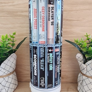 May include: A white tumbler with a blue background and a design of various cassette tapes featuring rock bands such as Iron Maiden, Slayer, Ozzy Osbourne, Judas Priest, Rush, Motley Crue, Celtic Frost, Nirvana, Scorpions, and Deep Purple.