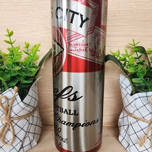 K&ouml;nnte beinhalten: Ein Edelstahlbecher mit einem rot-wei&szlig;en Budweiser-Etikett und dem aufgedruckten Text "City" und "Football Champions".