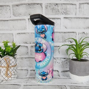 May include: A blue and pink stainless steel tumbler with a black lid and handle. The tumbler features a repeating pattern of Stitch from Lilo & Stitch holding a pink heart with the text "Ohana means family is love" in a circle around him.