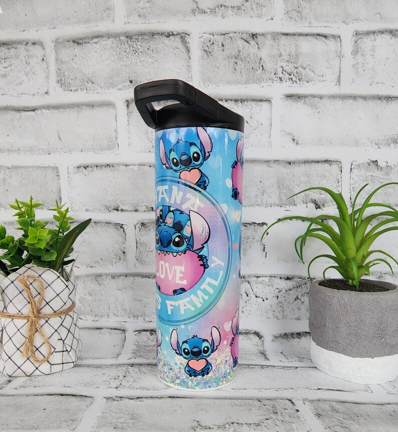 May include: A blue and pink stainless steel tumbler with a black lid and handle. The tumbler features a repeating pattern of Stitch from Lilo & Stitch holding a pink heart with the text "Ohana means family is love" in a circle around him.