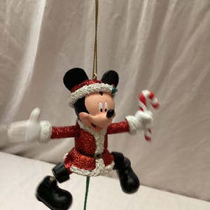 May include: A Christmas ornament featuring Mickey Mouse in a Santa-style outfit. Mickey is wearing a red glittery coat with white trim, black boots, and a Santa hat. He holds a candy cane and has a gold string for hanging.