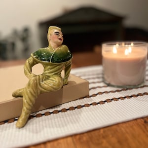May include: A ceramic figurine of a person wearing a green and yellow outfit sitting on a beige box. The figurine is on a white and brown striped surface.
