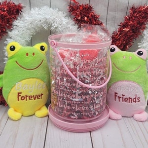 May include: Two green frog plush toys with embroidered names and messages. One says "Baylee Forever" and the other says "Blakelee Friends". A clear plastic jar with a pink lid and handle is filled with pink and silver glitter and hearts. The jar has the word "love" printed on it.