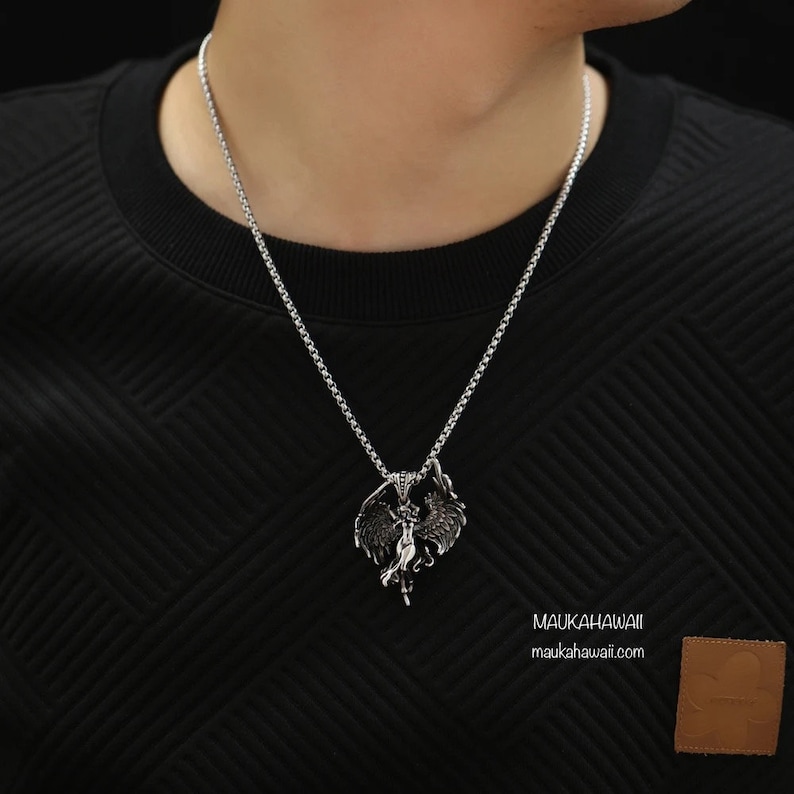 May include: A silver chain necklace with a pendant featuring a detailed design of a woman with wings. The pendant is made of metal and has a dark, aged finish. The necklace is worn by a person with a black shirt.