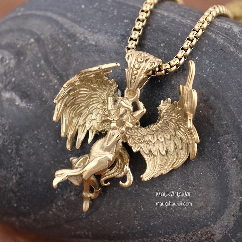 May include: Gold-toned pendant necklace featuring a detailed sculpture of a winged woman with outstretched wings. The pendant is attached to a gold chain.