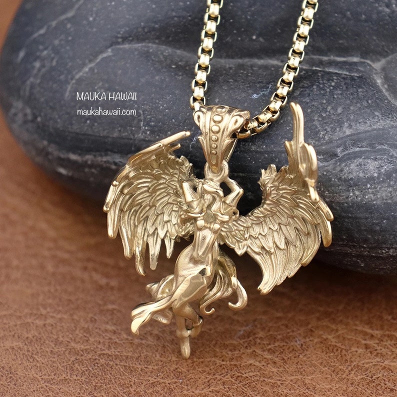 May include: Gold-toned pendant necklace featuring a detailed winged figure with a raised, textured design. The pendant is attached to a gold-toned chain.
