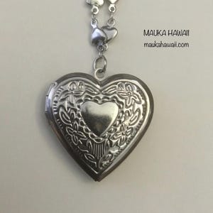 May include: Silver heart-shaped locket necklace with a floral design and a smaller heart in the center. The necklace is on a silver chain with heart-shaped links. The text "MAUKA HAWAII" and "maukahawaii.com" is printed on a white background.