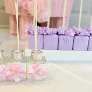 May include: Clear glass diffuser bottles with silver tops and pink flower accents. Each bottle has wooden reeds inserted. Behind the bottles are purple wrapped packages with decorative purple fabric. A pink basket with pink flowers is in the background.