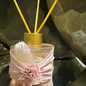 May include: A clear glass diffuser bottle with a gold lid and a pink fabric bow with a flower accent. The bottle is filled with a clear liquid and has several wooden sticks inserted into it.