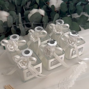 May include: Six small glass bottles with silver lids and white satin ribbon bows. Each bottle has a white flower and a label with text. The text on the labels reads "Hoşgeldiniz" and "Mutlu Yıllar".